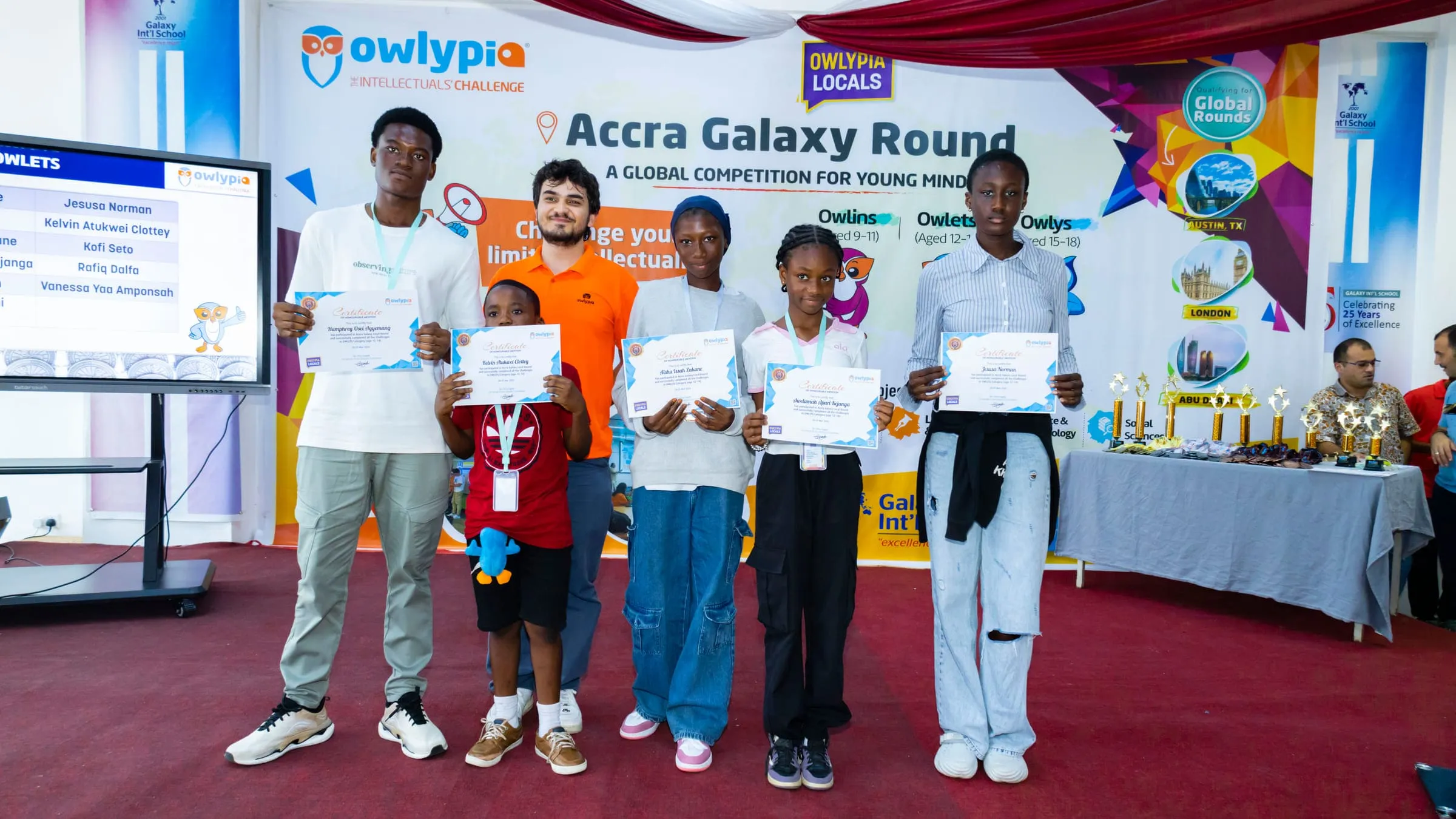 Pupils holding certificates in front of the Owlypia Accra Galaxy Round backdrop, trophies on a table behind