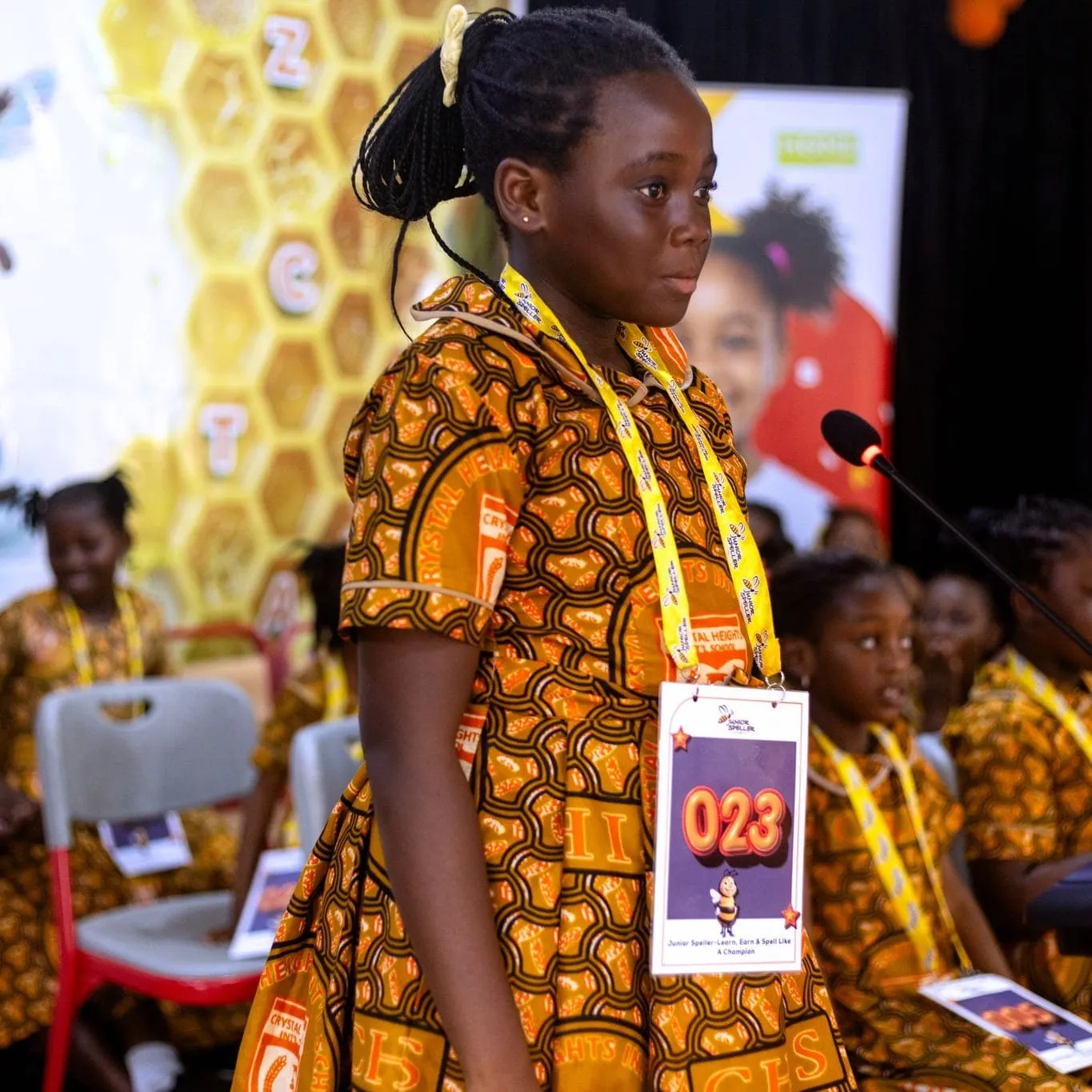 Crystal Heights pupils seated behind a contestant at the microphone during a spelling bee