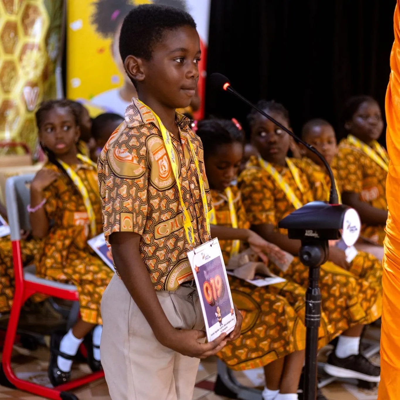 A Crystal Heights pupil at the microphone during a spelling bee, contestant number on a lanyard